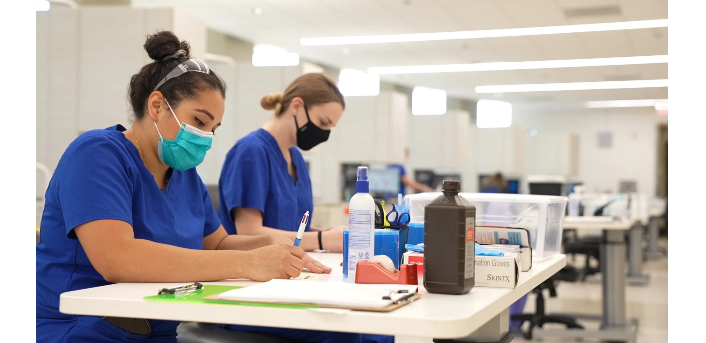 two female students wearing blue scrubs and sitting at lab table with books and pencils in hand