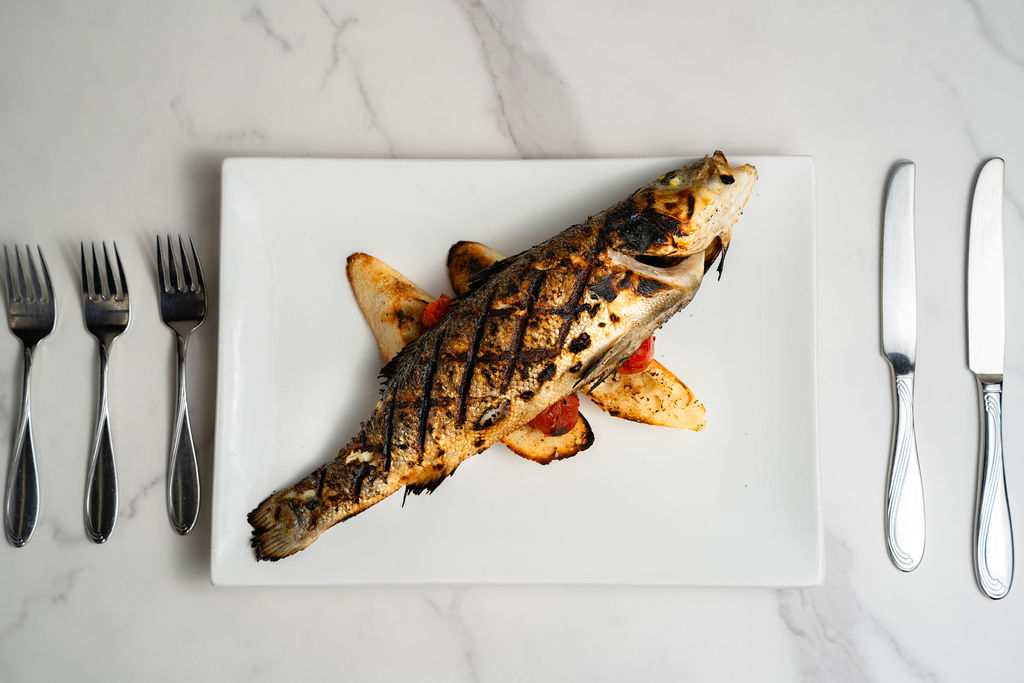 baked fish entree plated on white square plate, set on marble tabletop with three forks aligned to the left of the plate, and two knives aligned to the right of the plate.