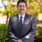 Dr. Roberto Gonzalez, the new president of Oxnard College, standing outdoors in a suit and tie, smiling with trees and greenery in the background.