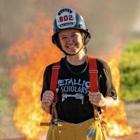 A young woman wearing a firefighter’s helmet and yellow pants stands smiling in front of a large fire. She has a black t-shirt with “Metallica Scholars” written on it. The background is an outdoor scene with flames visible behind her.