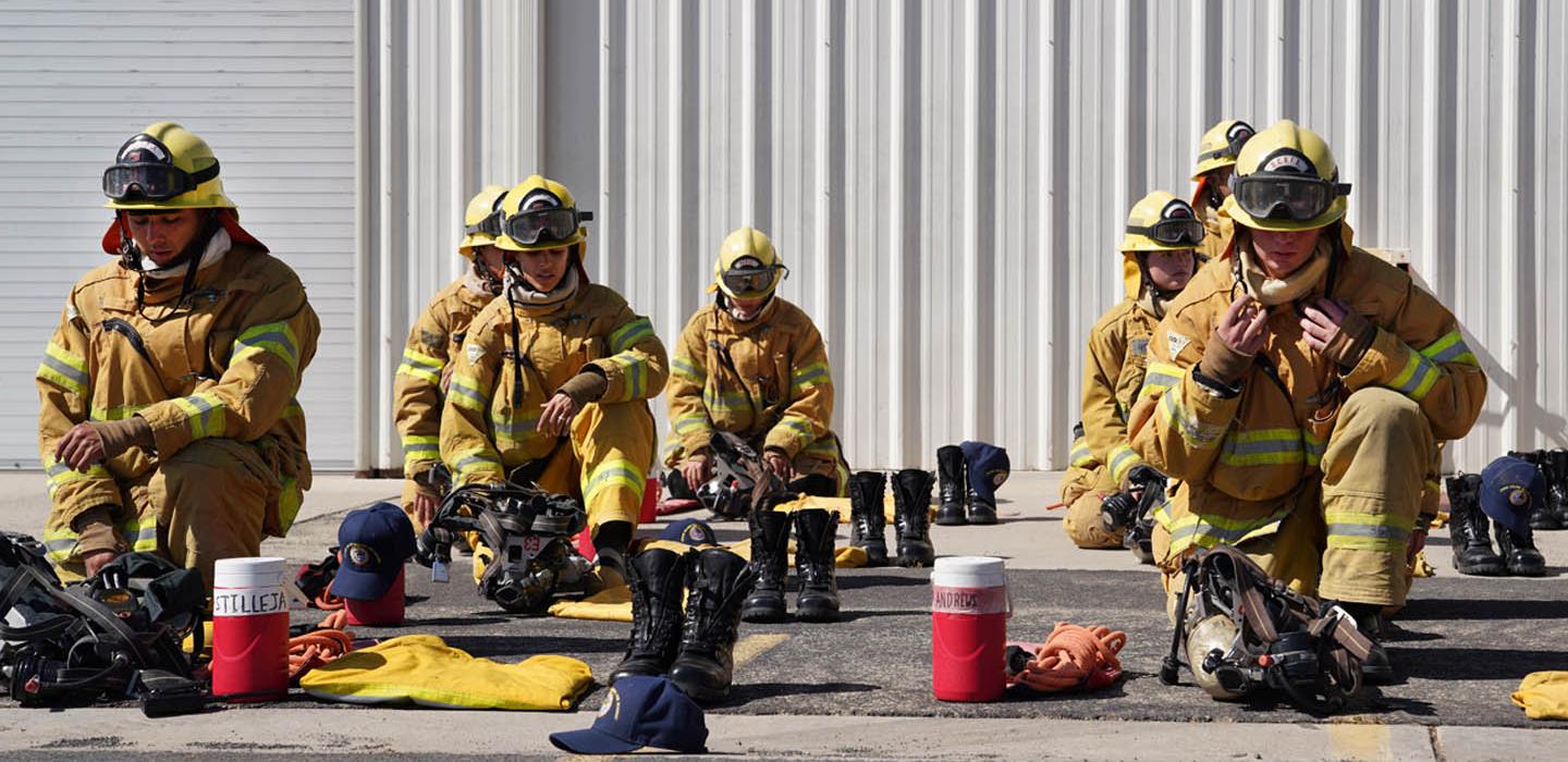Firefighters wearing yellow protective gear sit on the ground, arranging equipment in front of a white building with roll-up doors. Helmets, boots, and water jugs are scattered around them.