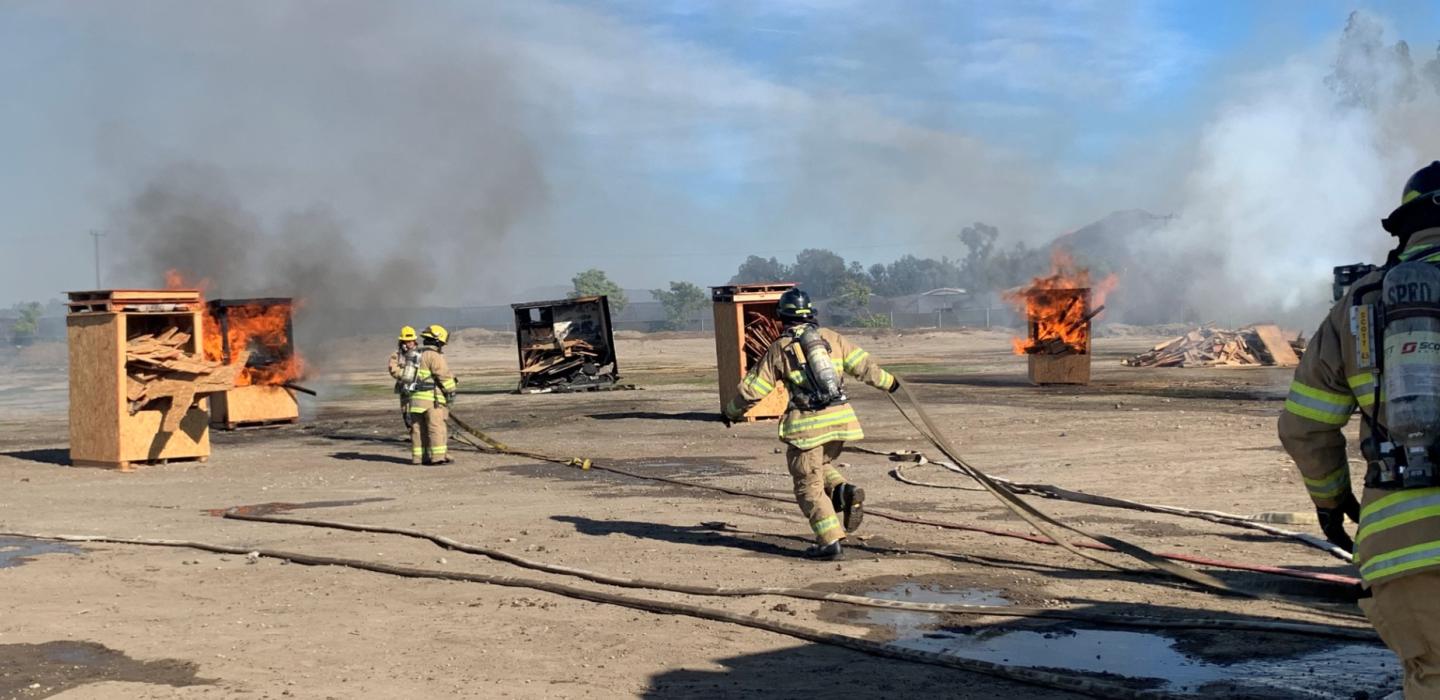 Firefighters extinguish burning wooden structures during a training exercise in an open field.