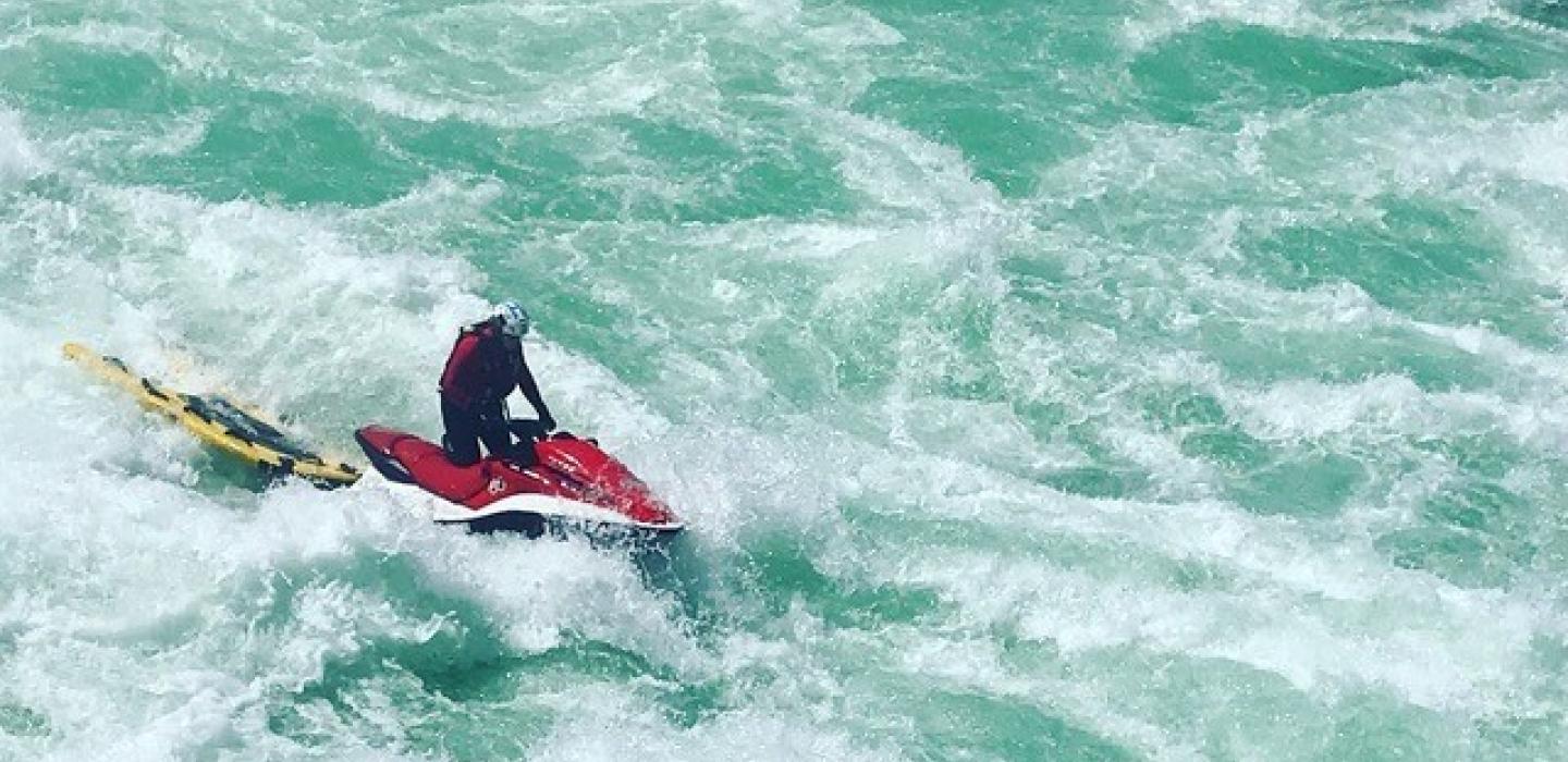 Person riding a red jet ski on turbulent, white-capped water.