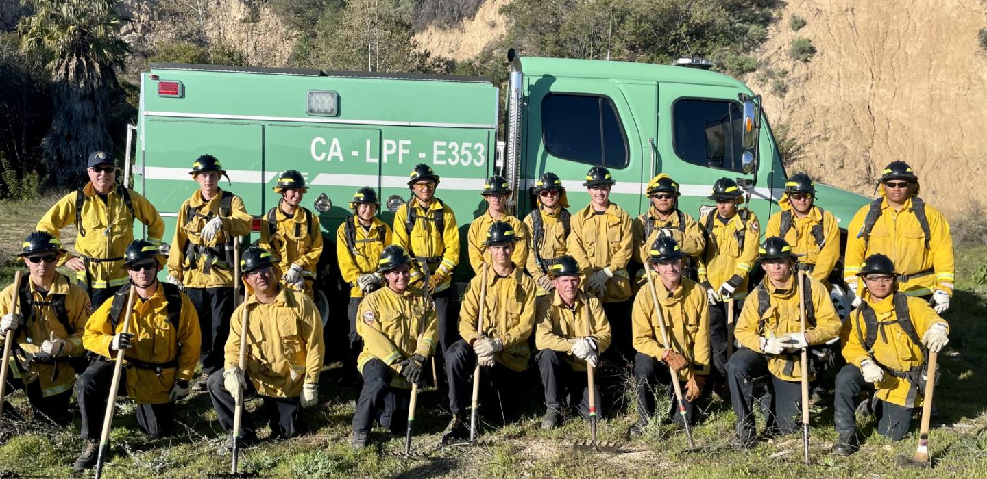 A group of firefighters in yellow uniforms and helmets pose in front of a green fire truck labeled "CA-LPF-E353" in an outdoor, grassy area with hills in the background.