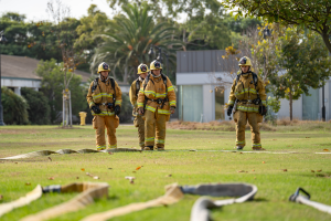 Four Fire Academy students standing outdoors in uniforms with a fire hose extended across the grass on the Oxnard College Public Safety Campus.