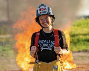 A young woman wearing a firefighter’s helmet and yellow pants stands smiling in front of a large fire. She has a black t-shirt with “Metallica Scholars” written on it. The background is an outdoor scene with flames visible behind her.